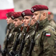 Żołnierze w bordowych beretach i mundurach polowych, z karabinami, reprezentują różne kategorie wojskowe. W tle biało-czerwona flaga.