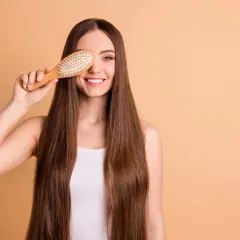 Mujer sonriente con pelo largo y liso, sosteniendo un cepillo de madera sobre su ojo. ¡Un truco para saber como desenredar un pelo muy enredado!