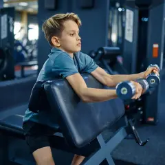Chłopiec ćwiczy biceps z hantlami na ławce. To jego siłownia dla 12 latka, gdzie buduje siłę.