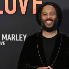 Uśmiechnięty mężczyzna w czapce i aksamitnej marynarce, przypominający Boba Marleya, z napisem "Bob Marley One Love" w tle. Może to być jeden z jego dzieci.