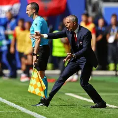 Sylvinho, trener w garniturze, gestykuluje na boisku. Obok stoi sędzia liniowy z flagą.