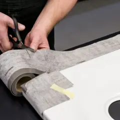 Hände schneiden Dichtungsband zu, um eine Duschwanne abzudichten. Das Material ist auf einer Rolle und wird präzise zugeschnitten.