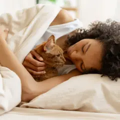 Frau mit Katze kuschelt im Bett unter einer Decke.