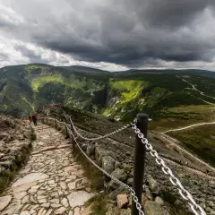 Kamienista ścieżka na szlak z kopy na Śnieżkę, z łańcuchami zabezpieczającymi. W oddali zielone zbocza i burzowe chmury.