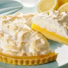 Pyszna tarta cytrynowa z bezą, z kawałkiem wyjętym na łopatce. Obok leżą świeże cytryny.