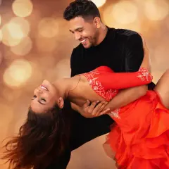 Coppia che balla con passione una salsa ballo latino, lei in abito rosso, lui in nero, su sfondo sfocato con luci dorate.