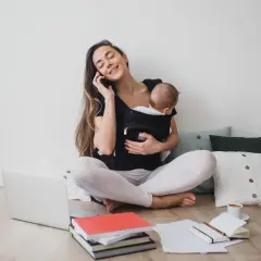 Femme au téléphone avec bébé en porte-bébé, entourée de livres et d'un ordinateur. Elle prépare sa lettre de prolongation de congé parental.