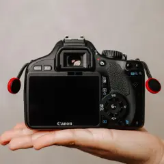 Dłoń trzyma czarną lustrzankę Canon z czerwonymi elementami smyczy.