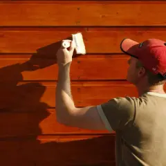 Un homme en casquette rouge est en train de peindre du bois brut extérieur avec un pinceau. La lumière du soleil projette une ombre nette sur le mur.