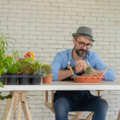 Mężczyzna z brodą i kapeluszem pochyla się nad stołem, otoczony roślinami. Czyżby miał alergię na kwiaty doniczkowe? Objawy mogą być różne.