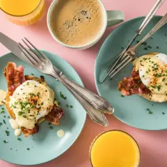 Zwei Teller mit **Egg Benedict**, garniert mit Speck und Schnittlauch, daneben Kaffee und Orangensaft.