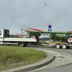 Transport przyczepy kempingowej na lawecie. Biała ciężarówka z platformą przewozi zielono-czerwoną przyczepę.