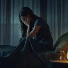 Mujer sentada en la cama, cubriéndose la cara con las manos, en plena vigilia del sueño. La noche es oscura, solo iluminada por una lámpara.