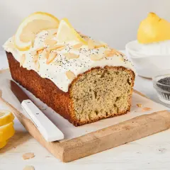 Ein saftiger Zitronen Mohn Kuchen mit weißem Zuckerguss, Mandelsplittern und Zitronenscheiben. Daneben liegen frische Zitronenscheiben und Mohn.