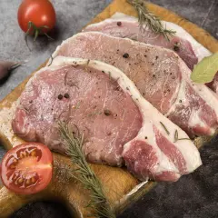 Surowe schaby na desce, przyprawione ziołami i pieprzem, gotowe do peklowania schabu do wędzenia. Obok pomidor, czosnek i liść laurowy.