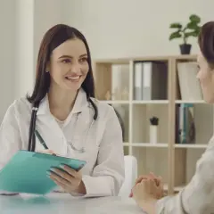 Lekarka w białym kitlu z receptą w ręku rozmawia z pacjentką w gabinecie. To może być wizyta w ramach programu AAP Norwegia.