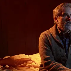 En man med grått skägg och glasögon sitter på en säng i ett dunkelt rum. Han ser bekymrad ut, som en patient som väntar på besked.
