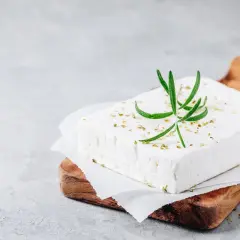 Un blocco di feta su un tagliere di legno, guarnito con rosmarino e spezie. Ricorda che la feta contiene lattosio.