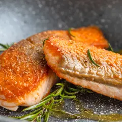 Zwei saftige Lachsfilets mit knuspriger Haut, perfekt gebraten, liegen in einer Pfanne mit Rosmarin und Öl.