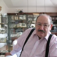 Wilhelm z Baskerville, w okularach i koszuli w paski, siedzi w otoczeniu antycznych ksiąg i gabinetów pełnych skarbów.