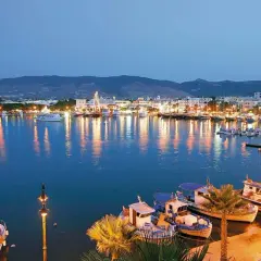 Hafen der Hauptstadt Kos bei Nacht, beleuchtete Stadt und Boote spiegeln sich im Wasser.