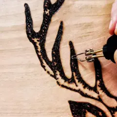 Dettaglio di lavori con pirografo su legno: una mano che disegna un palco di cervo con precisione.