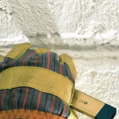 Mit einem Meißel wird alter **Putz entfernt**. Eine Hand mit Arbeitshandschuh hält das Werkzeug.