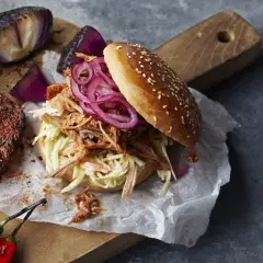 En saftig pulled pork-burgare med krispig coleslaw och rödlök, perfekt för ett bbq-recept. Bredvid ligger en bit grillat kött och rostade lökar.