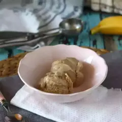 Zwei Kugeln cremiges, veganes Bananeneis in einer rosa Schale, daneben ein Löffel und ein Stück Gebäck. Eine Banane liegt im Hintergrund.