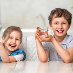 Uśmiechnięta dziewczynka i chłopiec trzymają szklanki z wodą. Cieszą się orzeźwiającym smakiem, jakby pili miękką wodę prosto ze źródła.