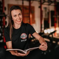 Trenerka z uśmiechem przegląda notatki, zastanawiając się, ile cardio na redukcji będzie najlepsze. W tle siłownia.