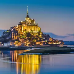 Mont Saint-Michel lyser upp i skymningen, en dröm för en semester i Frankrike. Vattnet speglar dess magiska ljus.