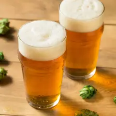 Dwa szklanki orzeźwiającego piwa hazy IPA z obfitą pianką, otoczone szyszkami chmielu na drewnianym blacie.