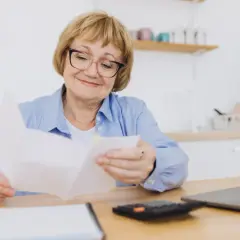 Uśmiechnięta starsza pani analizuje dokumenty, być może dotyczące ponownego przeliczenia emerytury ZUS. Obok leży kalkulator i laptop.
