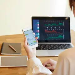 Frau analysiert Aktienkurse auf Laptop und Smartphone. Sie plant, wie sie ihre Aktiengewinne versteuern kann, während sie Notizen macht.