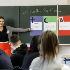 Lehrerin erklärt die Goldene Regel im Religionsunterricht Grundschule. Tafeln zeigen Zitate aus Christentum, Islam, Alevitentum und Buddhismus.