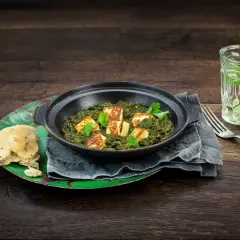 En skål med krämig palak paneer, toppad med gyllene paneerbitar och färsk koriander. Serveras med naanbröd och en glasdryck.