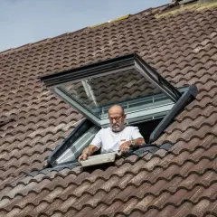 Mężczyzna montuje okno dachowe balkonowe na brązowym, dachówkowym dachu.