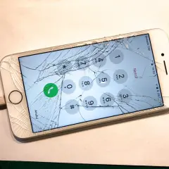 Zepsuty ekran telefonu z widocznym pęknięciem, podłączony do ładowarki. Na ekranie widoczna klawiatura numeryczna.