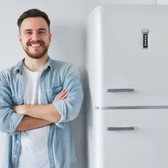 Uśmiechnięty mężczyzna w jeansowej koszuli stoi obok białej lodówki.
