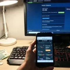 Dłoń trzyma smartfon z otwartą stroną Skinport, obok klawiatury i pieniędzy.