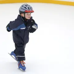 Bambino con casco e tuta blu pratica il pattinaggio sul ghiaccio, con i pattini azzurri e lacci arancioni.