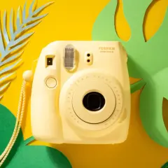 Żółty aparat natychmiastowy Fujifilm Instax Mini 8 na tle żółtym z zielonymi i niebieskimi liśćmi. Idealny do wakacyjnych zdjęć, często pojawia się w rankingach aparatów natychmiastowych.