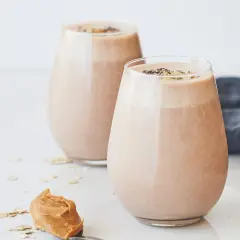 Två glas med krämig choklad- och jordnötssmoothie, toppad med chiafrön. En sked med jordnötssmör och havregryn ligger bredvid. En perfekt start på dagen, som Don Pedro skulle ha gillat.