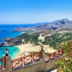 Blick auf eine Bucht mit türkisfarbenem Wasser, Sandstrand und grünen Hügeln. Ein Geländer mit Laternen und blühenden Oleandern im Vordergrund. Rhodos Geheimtipps für unvergessliche Ausblicke.