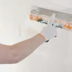 Mano con guanto bianco stende stucco murale su una parete bianca con una spatola arancione.