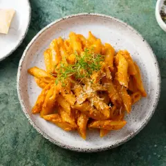Penne z aromatycznym, czerwonym pesto, posypane parmezanem i świeżymi ziołami. Obok miseczka z solą i pieprzem.