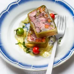 Deux portions de fromage de tête translucide, garnies de légumes et de viande, sur une assiette ornée de bleu.
