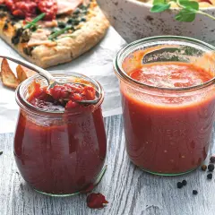 Dwa słoiki domowego sosu do pizzy, jeden z łyżką pełną aromatycznej masy. Obok kawałek pizzy i przyprawy.