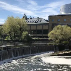 Vattenfall porlar förbi Louis de Geer Konsert & Kongress, en modern byggnad med en rundad takkonstruktion, omgiven av grönska och gamla industribyggnader.